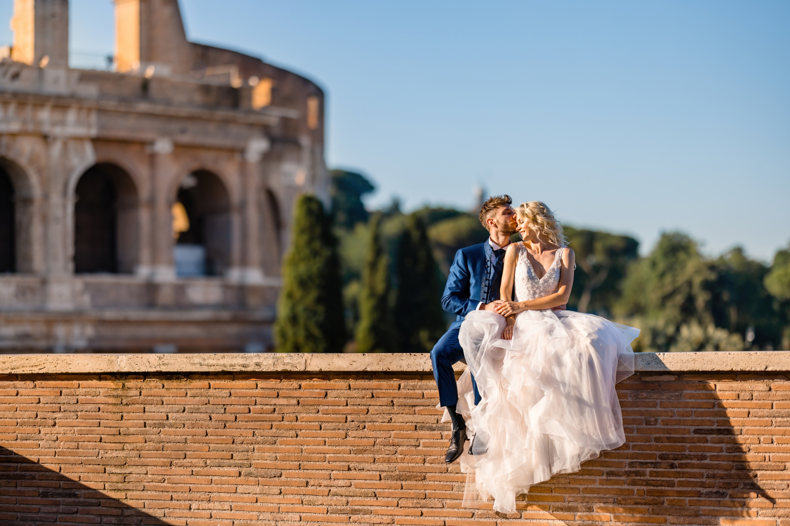 bride and groom sitting on a wall in front of colosseum in Rome where the brides puffy dress hangs beautifully and photographed by destination wedding photographer