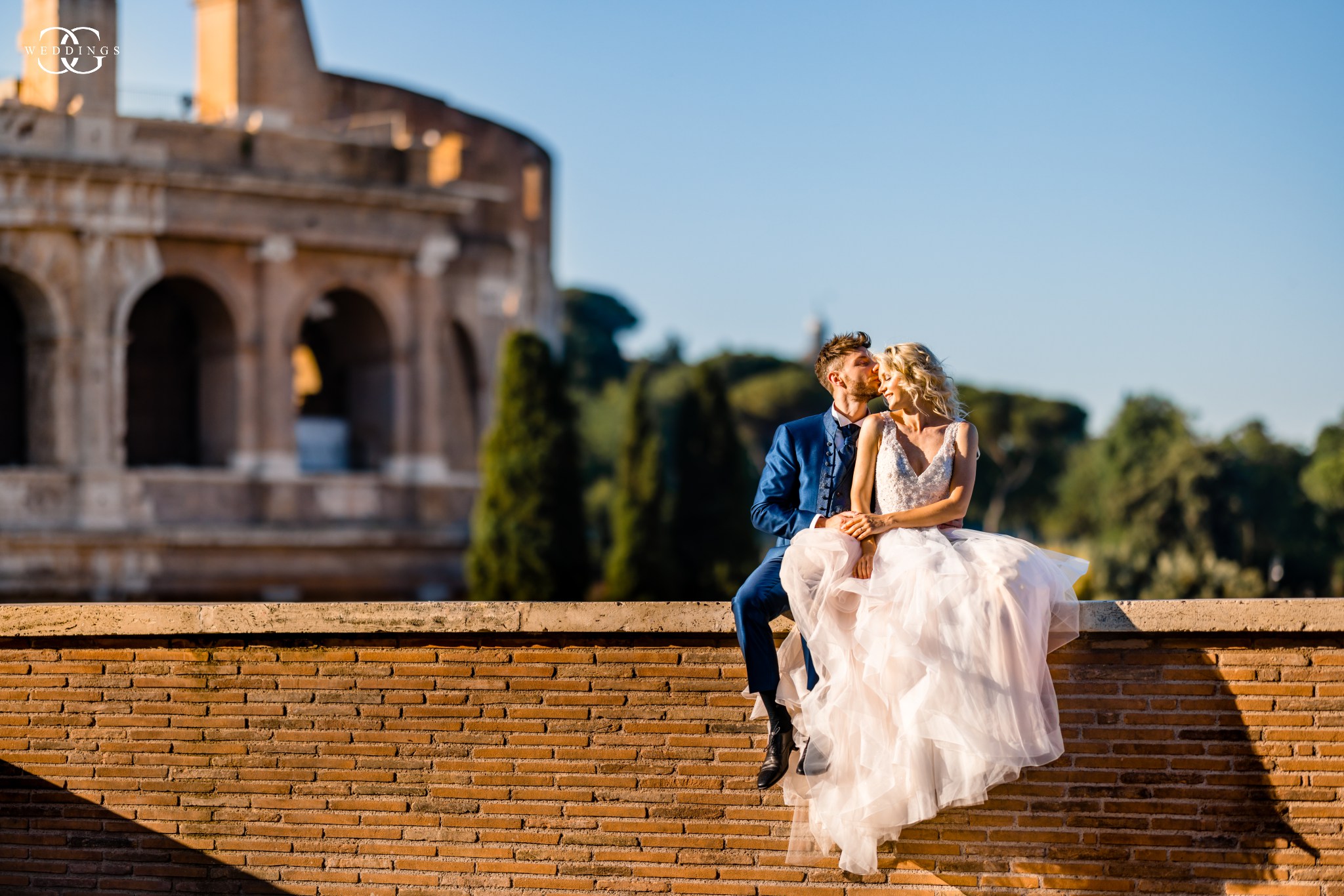 bride and groom sitting on a wall in front of colosseum in Rome where the brides puffy dress hangs beautifully and photographed by destination wedding photographer
