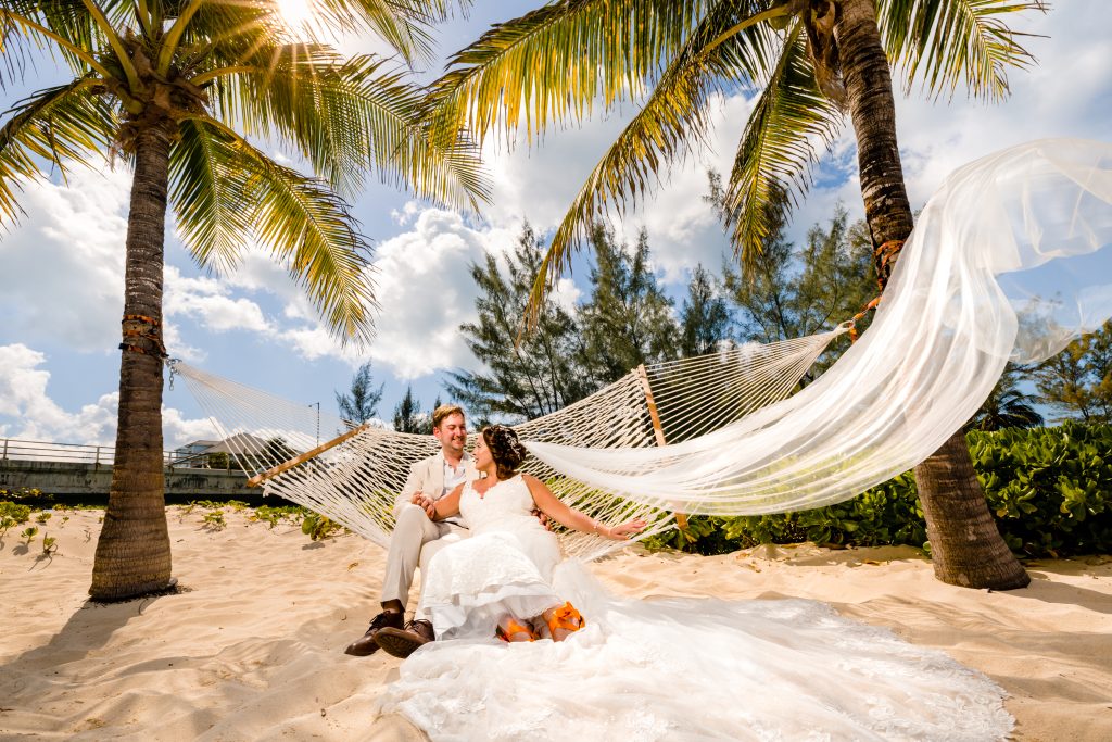 bride and groom sitting in a hammock and holding hands for their wedding photo while the wind blows her veil away.