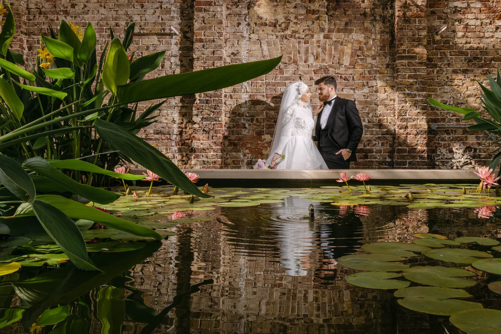 Bride and groom beside lily pond and brick wall