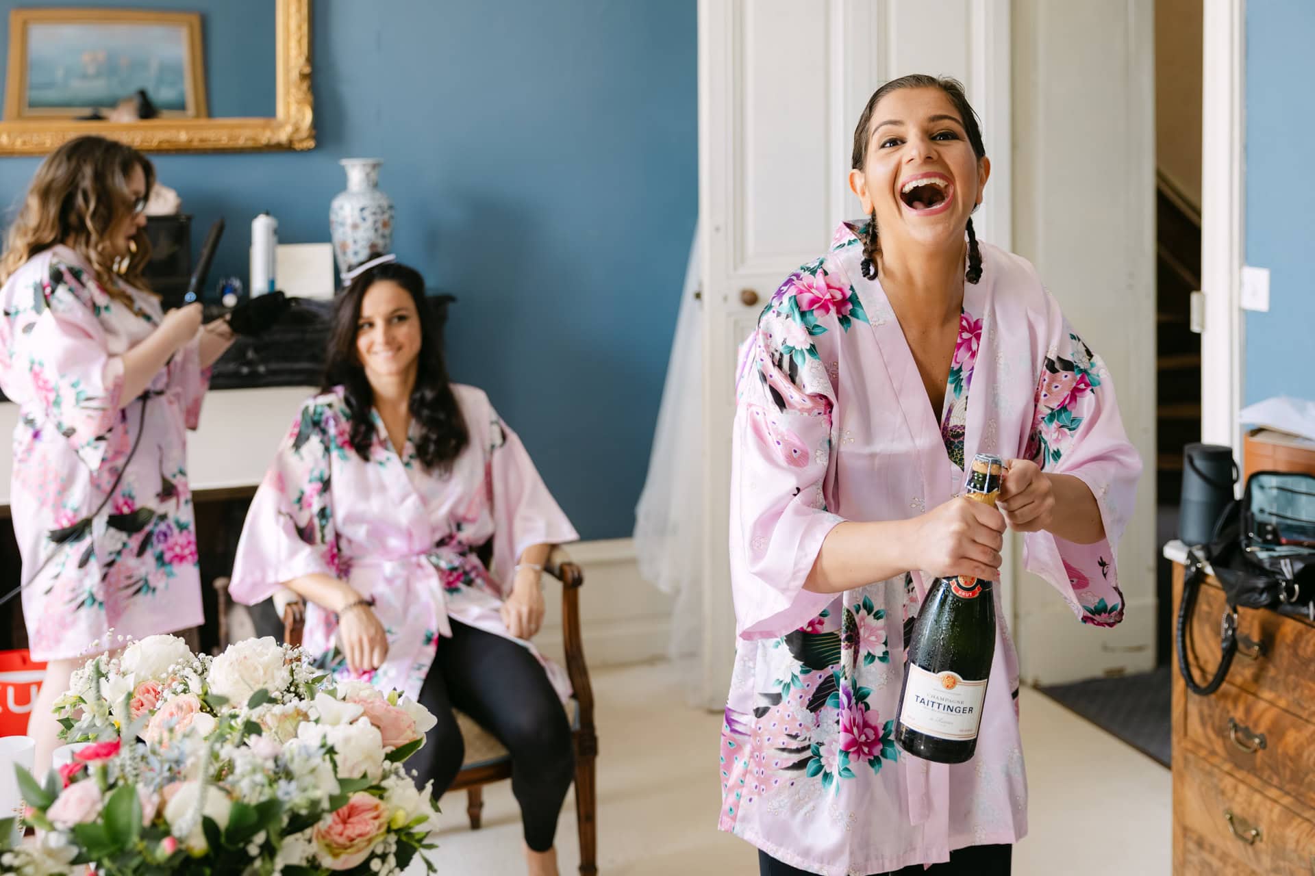 Bridesmaids in floral robes opening champagne indoors